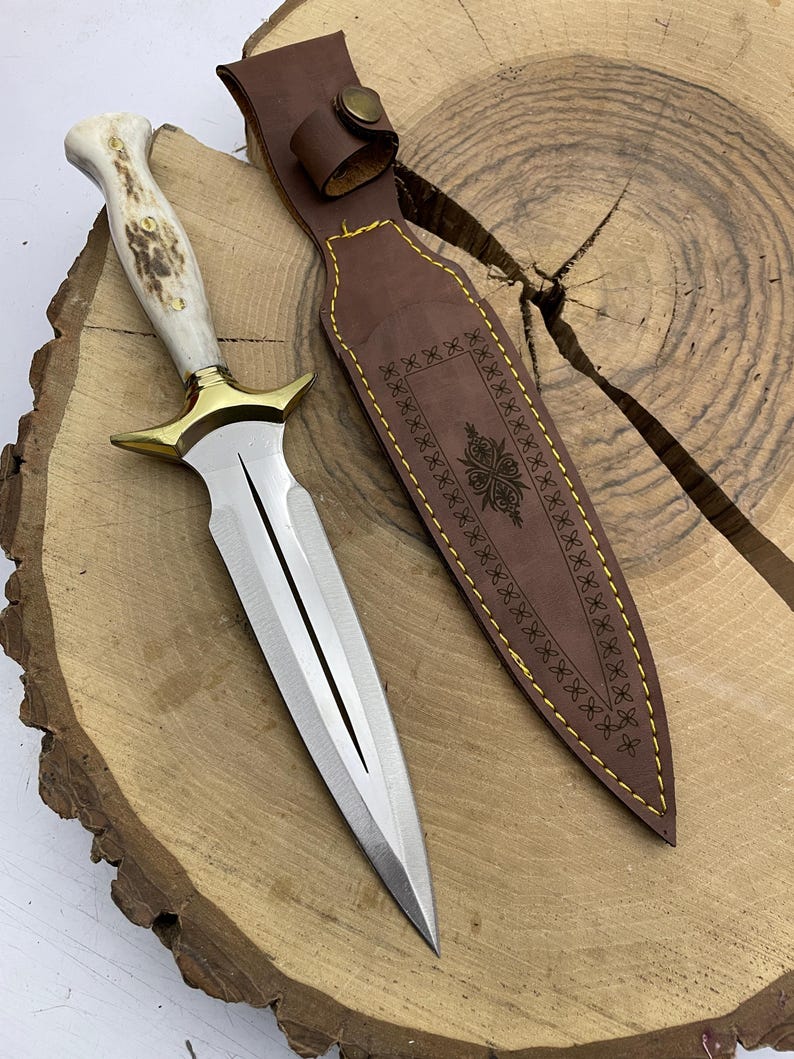 May include: A silver dagger with a white antler handle and a gold-coloured guard. The dagger is on a wooden surface and has a brown leather sheath with decorative stitching.