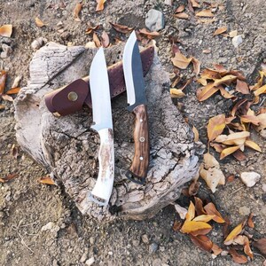 May include: Two hunting knives with wooden and bone handles rest on a weathered tree stump. One knife has a light-coloured bone handle, while the other has a dark wood handle. A brown leather sheath is visible behind the knives.