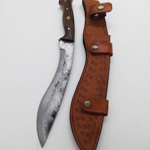 May include: A curved, silver-colored knife with a wooden handle and a brown leather sheath. The sheath has a decorative pattern and a snap closure.