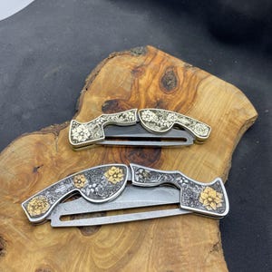 Peut inclure: Deux couteaux pliants avec des motifs floraux. Les manches pr&eacute;sentent des motifs floraux argent&eacute;s et dor&eacute;s complexes. Les lames sont argent&eacute;es. Les couteaux sont expos&eacute;s sur une surface en bois.