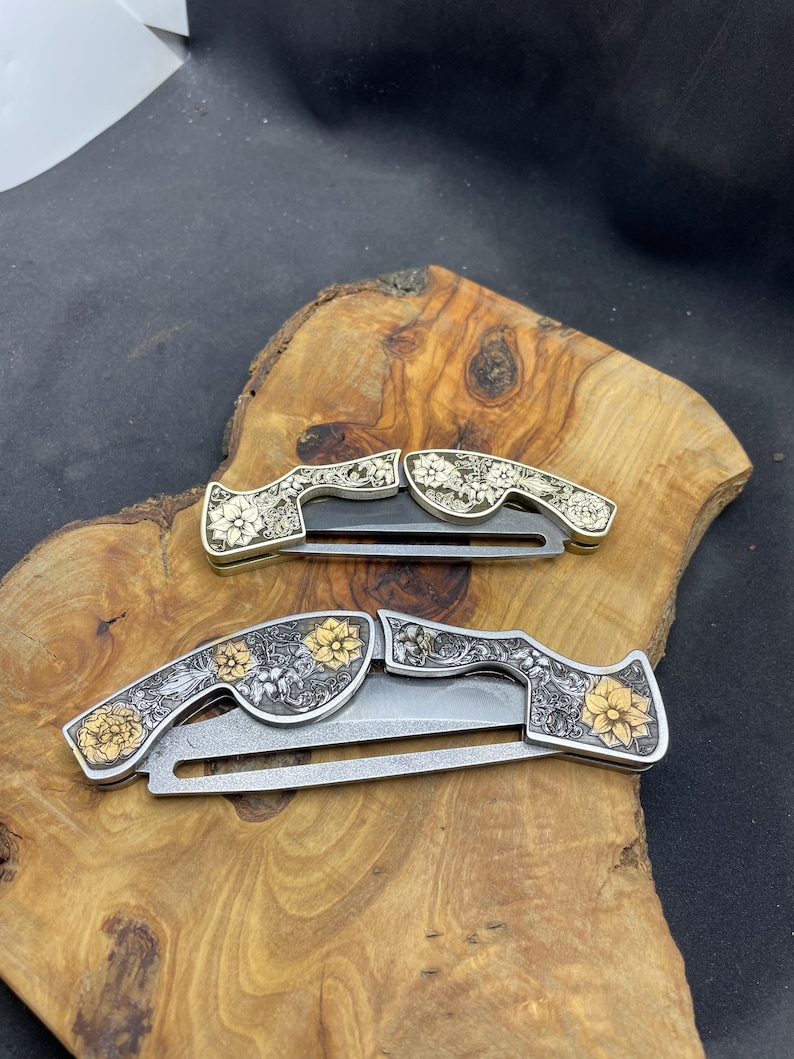 Peut inclure: Deux couteaux pliants avec des motifs floraux. Les manches pr&eacute;sentent des motifs floraux argent&eacute;s et dor&eacute;s complexes. Les lames sont argent&eacute;es. Les couteaux sont expos&eacute;s sur une surface en bois.