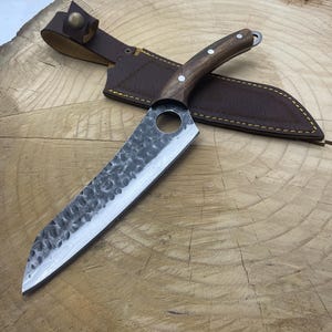 May include: A black and silver chef's knife with a wooden handle and a leather sheath. The knife has a hammered finish and a round hole in the blade.