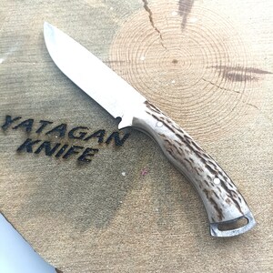 May include: A stainless steel knife with a light brown and white handle. The knife blade is silver and the handle has a textured pattern. The text "YATAGAN KNIFE" is visible on the wooden surface.