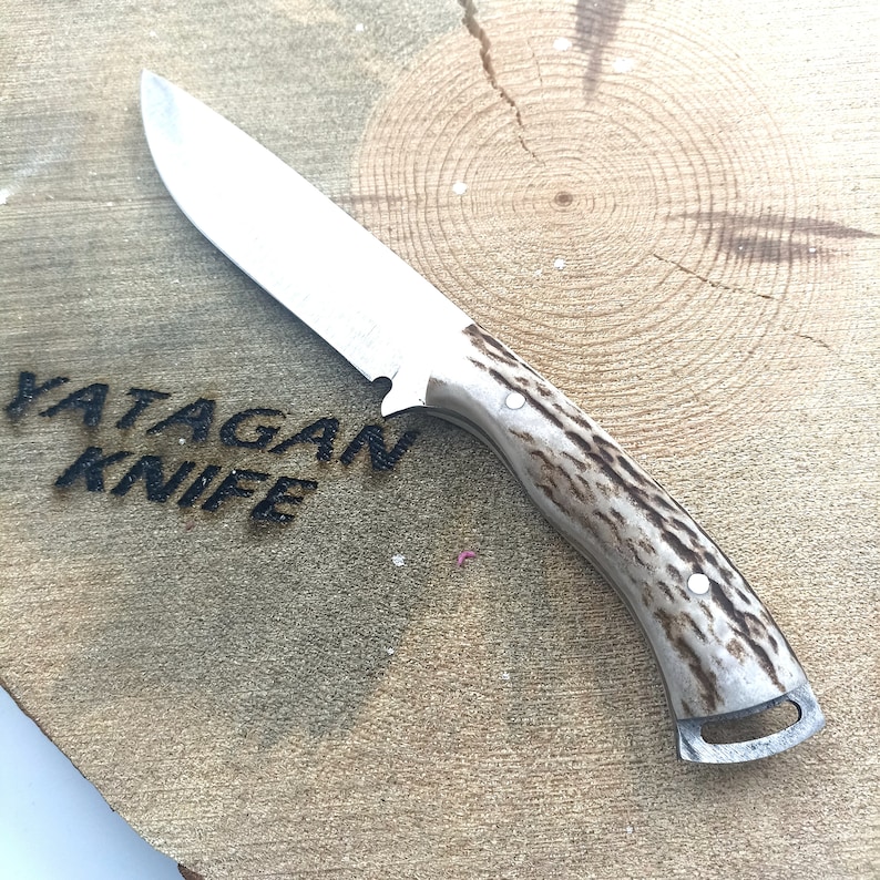 May include: A stainless steel knife with a light brown and white handle. The knife blade is silver and the handle has a textured pattern. The text "YATAGAN KNIFE" is visible on the wooden surface.