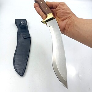 Puede incluir: Un cuchillo plateado con mango de madera marr&oacute;n y una funda de cuero negro. El cuchillo tiene una hoja curva y una guarda de lat&oacute;n.