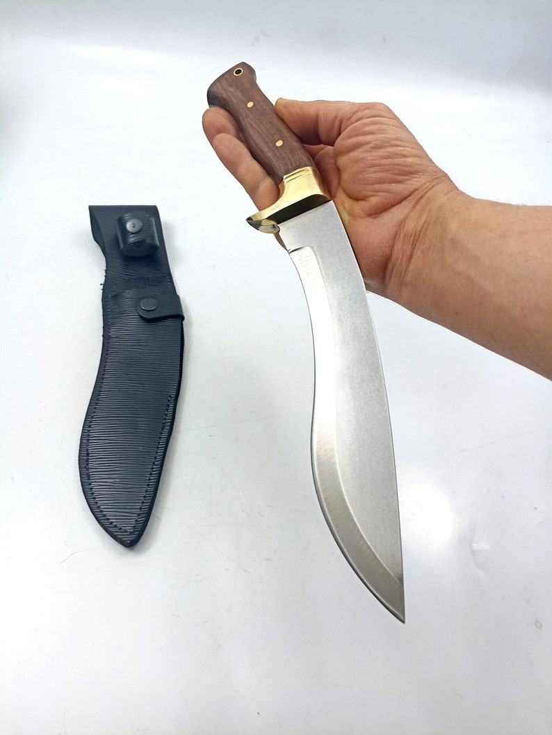 Puede incluir: Un cuchillo plateado con mango de madera marr&oacute;n y una funda de cuero negro. El cuchillo tiene una hoja curva y una guarda de lat&oacute;n.