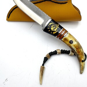 May include: A small, silver knife with a black handle and a wolf design. The handle is made of bone and has a brown and gold inlay. The knife comes with a brown leather sheath and a black cord with bone beads.