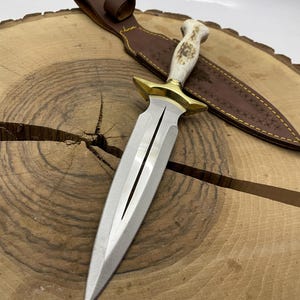 May include: A silver dagger with a white bone handle and gold accents. The dagger is on a wooden surface and has a brown leather sheath with decorative stitching.
