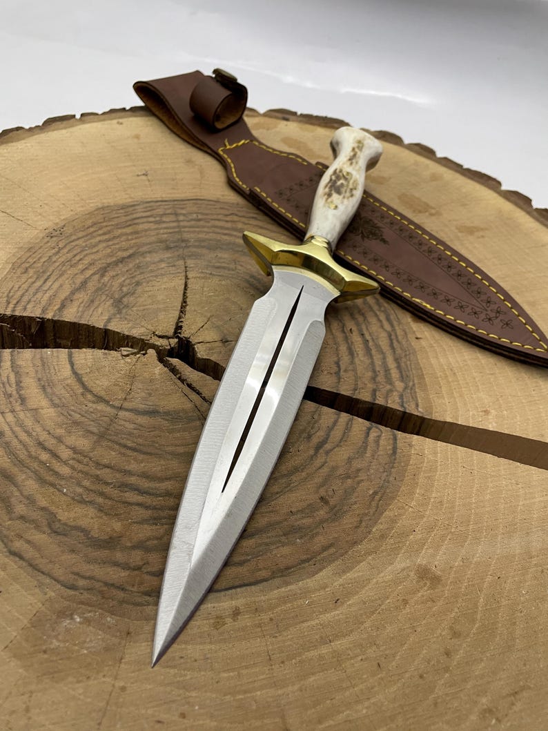 May include: A silver dagger with a white bone handle and gold accents. The dagger is on a wooden surface and has a brown leather sheath with decorative stitching.