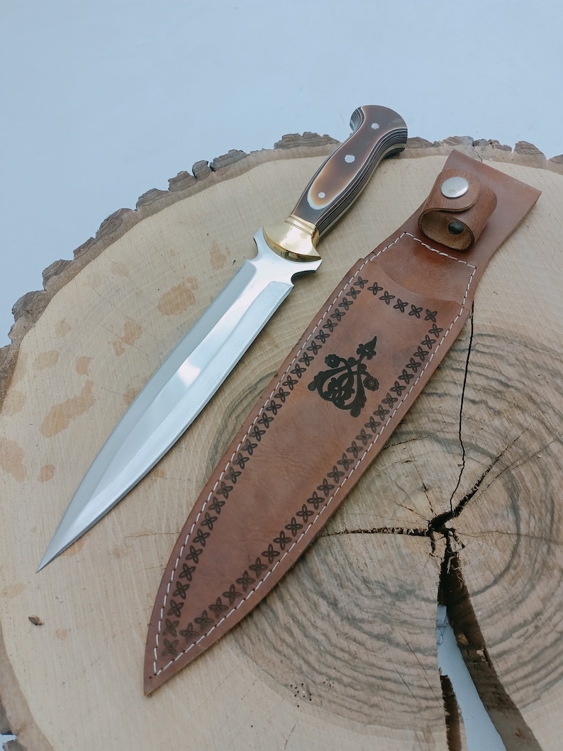 Op de afbeelding: Een bruine leren schede met een decoratief ontwerp en een zilveren dolk met een bruin en wit handvat.