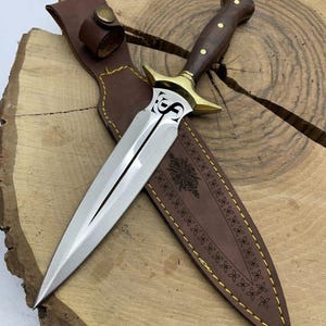 Op de afbeelding: Een zilveren dolk met een bruin houten handvat en een gouden beschermkap. De dolk zit in een bruine leren schede met een decoratief patroon.