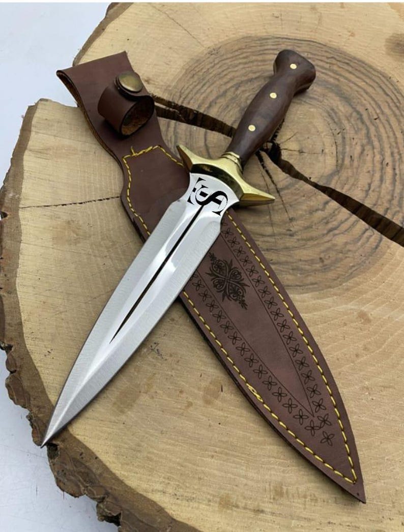May include: A silver dagger with a brown wooden handle and a gold-coloured guard. The dagger is in a brown leather sheath with a decorative pattern.