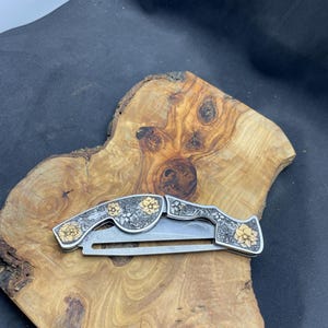 Peut inclure: Un couteau pliant argent&eacute; avec des motifs floraux dor&eacute;s. Le couteau est ouvert, r&eacute;v&eacute;lant la lame. Le manche et l'extr&eacute;mit&eacute; du couteau sont orn&eacute;s de fleurs dor&eacute;es et de motifs complexes. Le couteau repose sur une surface en bois.