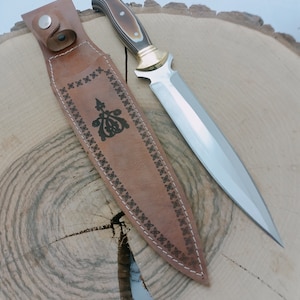 Op de afbeelding: Een bruine leren schede met ingewikkelde stiksels en een zilveren mes met een bruin en zwart handvat.
