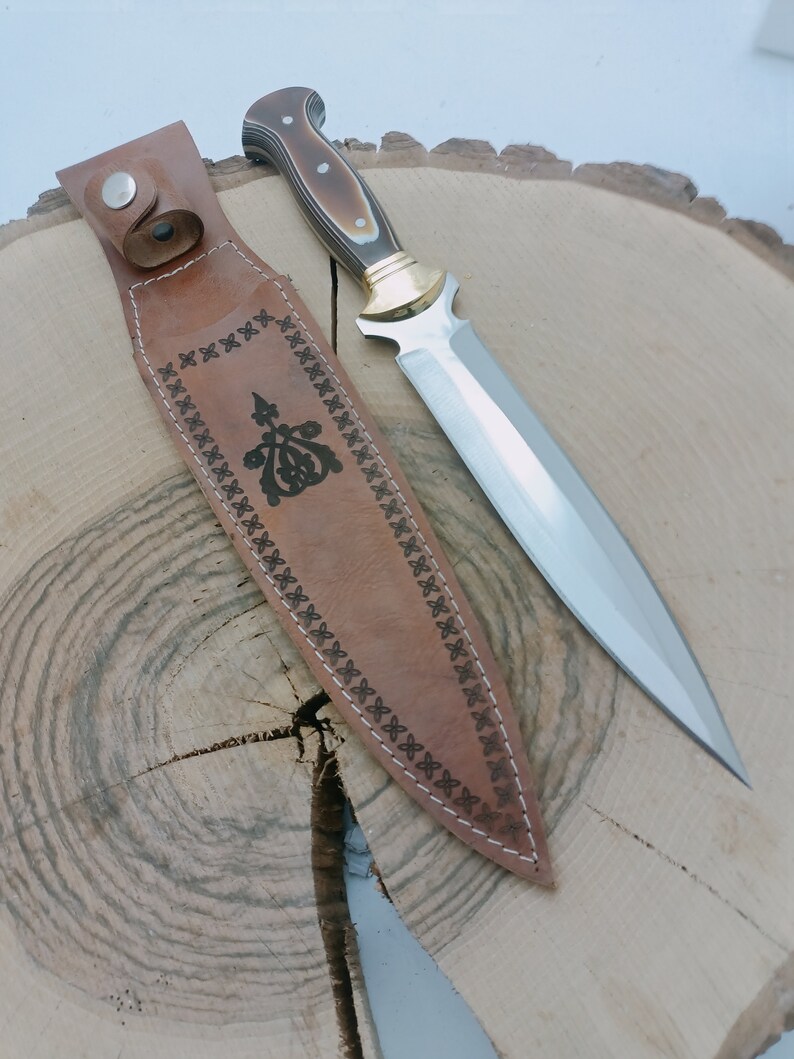 Op de afbeelding: Een bruine leren schede met ingewikkelde stiksels en een zilveren mes met een bruin en zwart handvat.