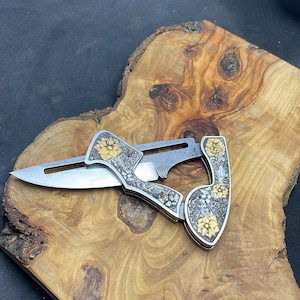 Peut inclure: Un couteau pliant argent&eacute; avec un motif floral et des accents dor&eacute;s. Le couteau est ouvert, r&eacute;v&eacute;lant une lame tranchante. La poign&eacute;e pr&eacute;sente des gravures complexes de fleurs et de feuilles. Le couteau est pr&eacute;sent&eacute; sur une surface en bois.