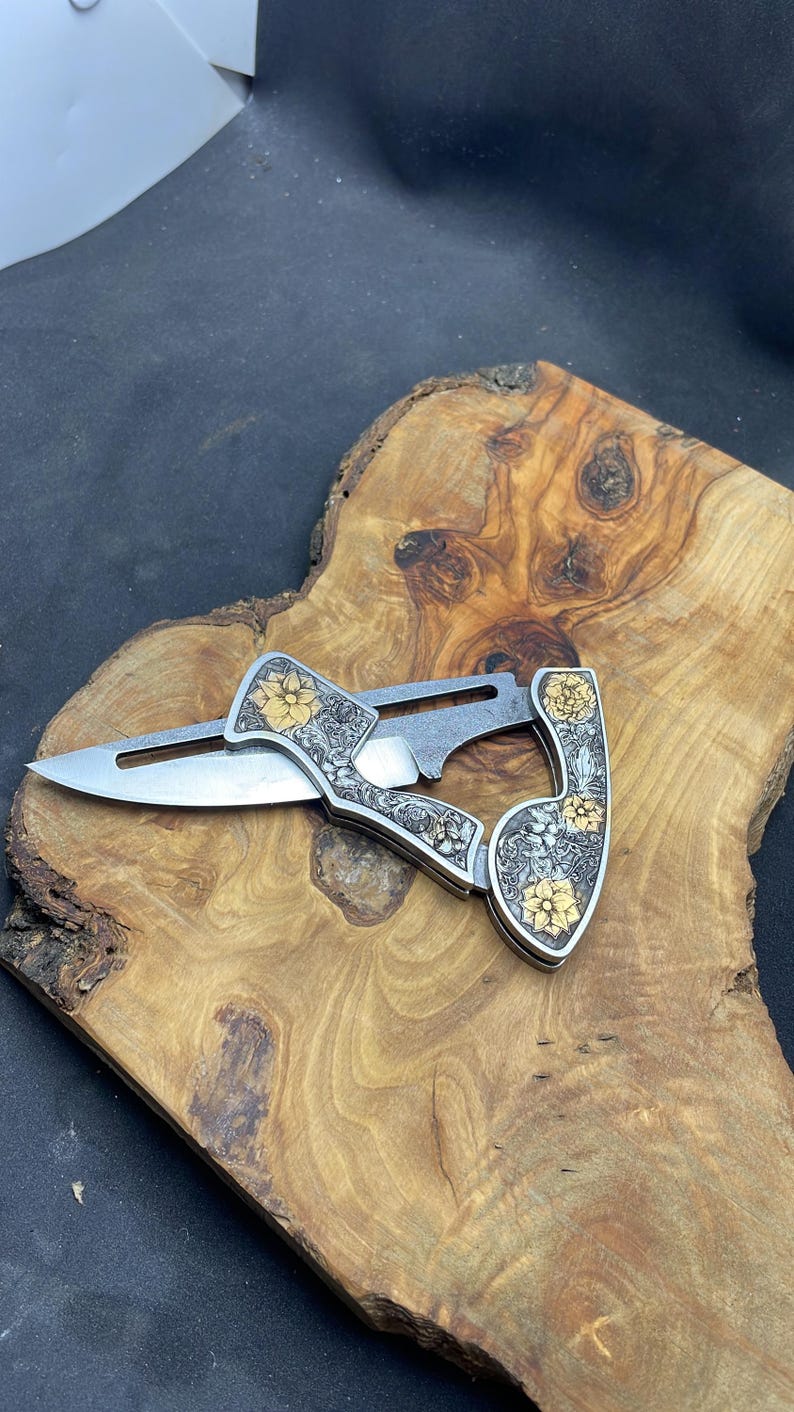 Peut inclure: Un couteau pliant argent&eacute; avec un motif floral et des accents dor&eacute;s. Le couteau est ouvert, r&eacute;v&eacute;lant une lame tranchante. La poign&eacute;e pr&eacute;sente des gravures complexes de fleurs et de feuilles. Le couteau est pr&eacute;sent&eacute; sur une surface en bois.