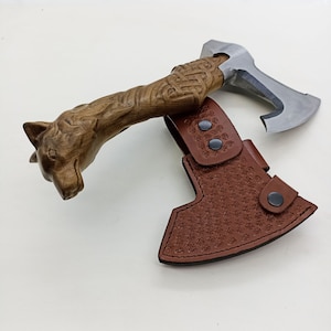 May include: A brown leather axe sheath with a wolf head handle. The sheath has a detailed pattern and a snap closure.
