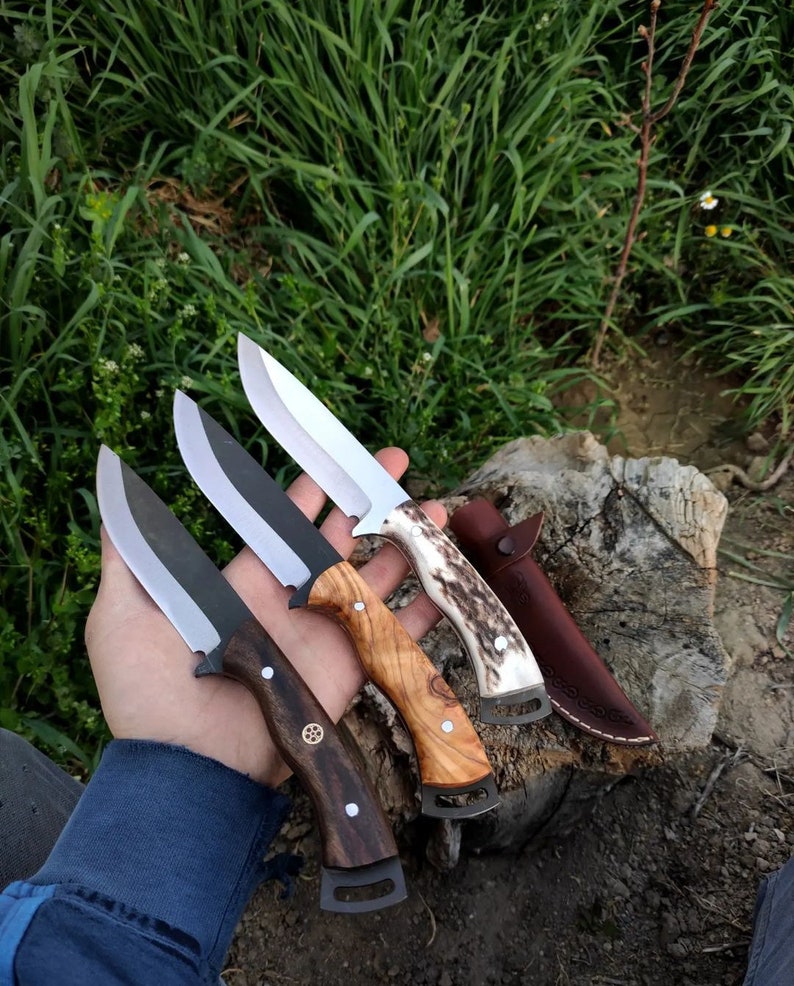 May include: Four custom hunting knives with diverse handle materials, including wood and antler, and a leather sheath. The blades are silver and black, and the handles display unique grain patterns and decorative elements. The knives are displayed outdoors.