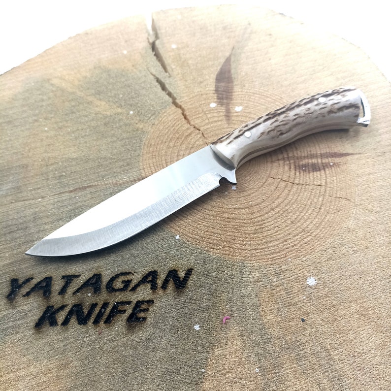 May include: A stainless steel knife with a light-coloured, textured handle rests on a wooden surface. The knife has a long, sharp blade and the text "YATAGAN KNIFE" is carved into the wood.