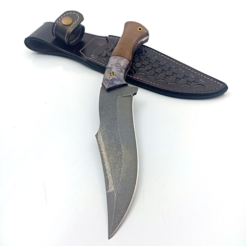 May include: A black and grey knife with a brown and purple handle. The knife is in a brown leather sheath with a decorative pattern.