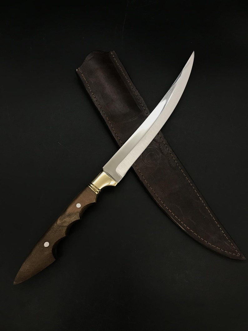 May include: A long, curved, silver knife with a brown wooden handle and a leather sheath. The knife is lying on a black surface.