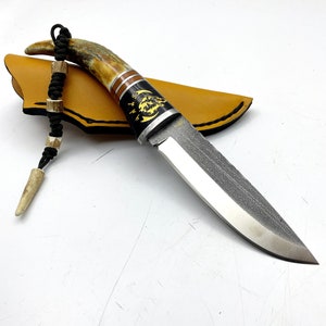 May include: A hunting knife with a brown and black handle and a black leather sheath. The handle is decorated with a wolf design and a moon.
