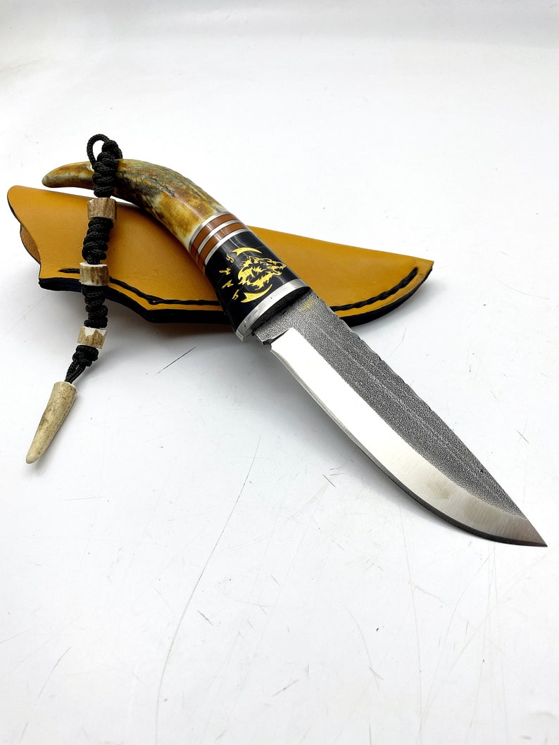 May include: A hunting knife with a brown and black handle and a black leather sheath. The handle is decorated with a wolf design and a moon.