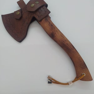 May include: A handcrafted axe with a wooden handle and a brown leather sheath. The axe head is made of metal and has a sharp edge. The leather sheath is secured with brass rivets and features decorative tooling. A lanyard is attached to the handle.