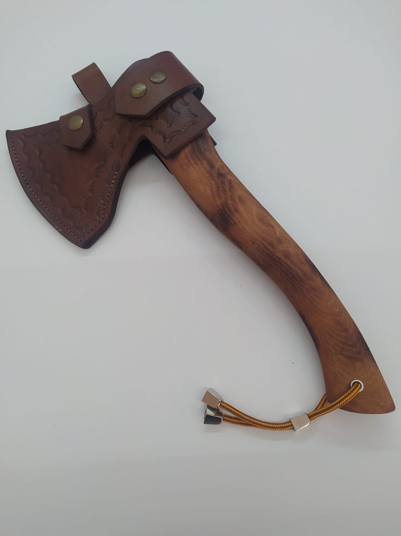 May include: A handcrafted axe with a wooden handle and a brown leather sheath. The axe head is made of metal and has a sharp edge. The leather sheath is secured with brass rivets and features decorative tooling. A lanyard is attached to the handle.