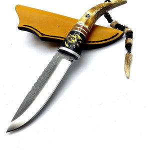 May include: A hunting knife with a black handle and a brown leather sheath. The handle is made of antler and has a decorative design. The blade is serrated and has a polished finish.