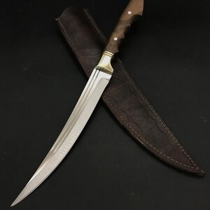 May include: A curved, silver-colored knife with a wooden handle and a leather sheath. The handle has a brass accent and two white dots.