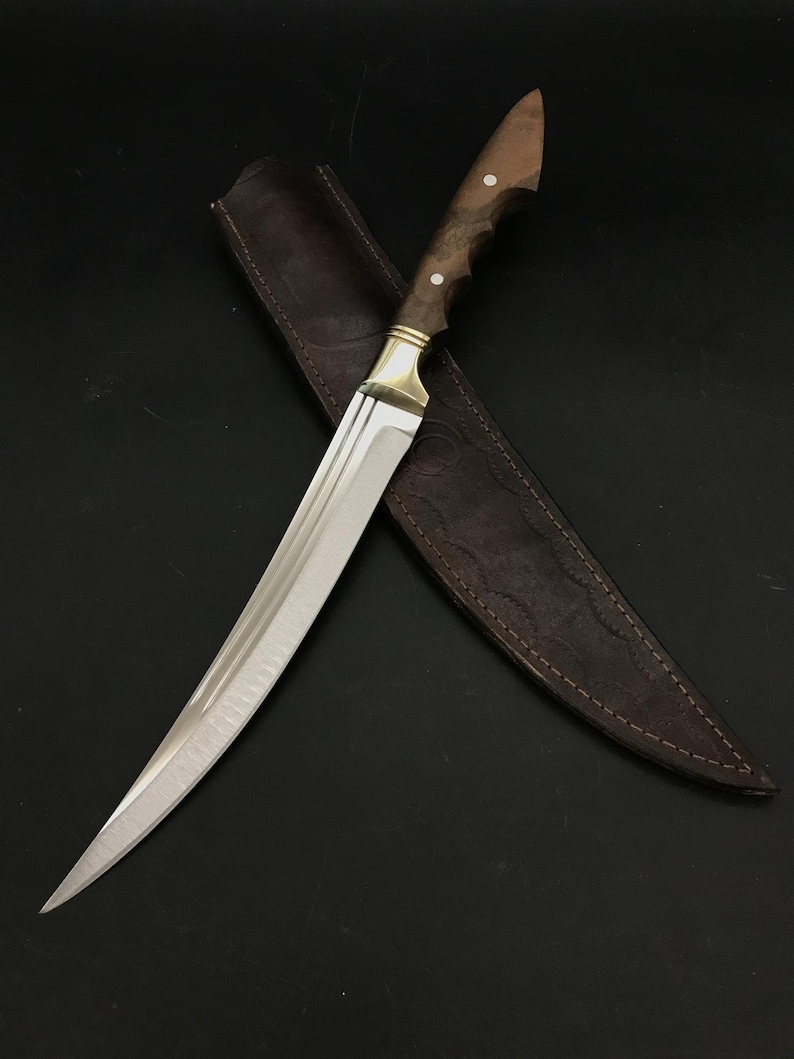 May include: A curved, silver-colored knife with a wooden handle and a leather sheath. The handle has a brass accent and two white dots.