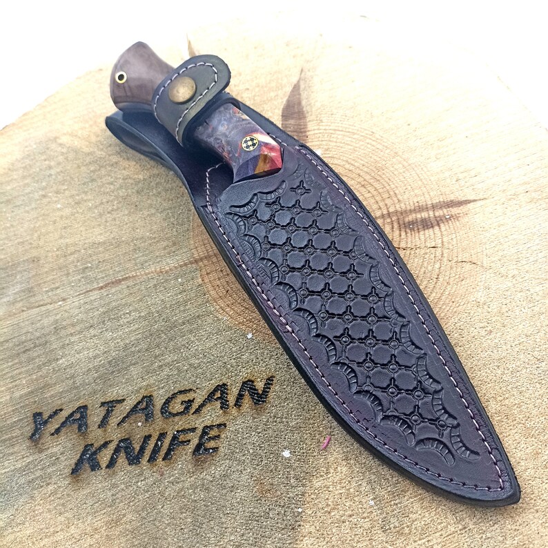May include: A brown leather sheath with a decorative pattern and a stitched edge. A knife with a wooden handle and a multi-colored blade is inside the sheath. The text "YATAGAN KNIFE" is burned into the wooden surface the sheath is resting on.