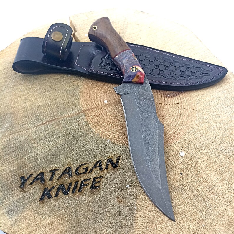 May include: A black and grey YATAGAN KNIFE with a wooden handle and a leather sheath. The knife has a curved blade and a textured finish.