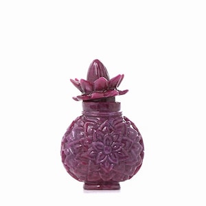 Puede incluir: Un frasco de perfume de color magenta oscuro con un cuerpo en forma de piña y un tapón en forma de flor. La botella presenta patrones florales tallados y se encuentra sobre un fondo blanco. El diseño es intrincado y elegante.