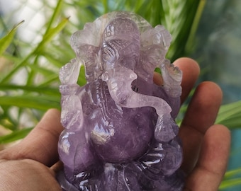 Puede incluir: Una pequeña estatua de Ganesha de amatista púrpura tallada con detalle. La estatua está sentada sobre una base y tiene la trompa levantada en un gesto de bendición.