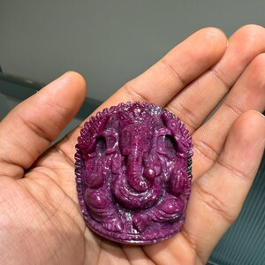 May include: A carved, deep magenta gemstone pendant depicting the Hindu deity Ganesha. The pendant features intricate details of the elephant-headed god in a seated pose. The gemstone is held in a hand, showcasing its rich colour and detailed carving.