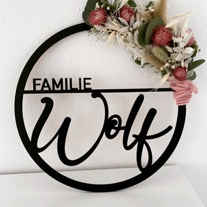 Könnte beinhalten: Schwarzer Holz-Kreis mit dem Schriftzug "Familie Wolf" in Kursivschrift. Der Kreis ist mit getrockneten Blumen und Grünpflanzen verziert.