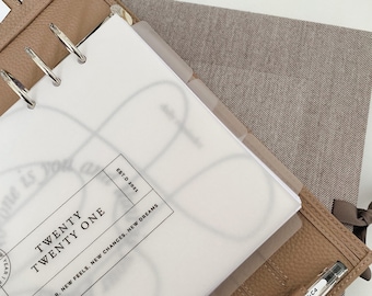 May include: A tan leather planner with silver rings, open to a page that reads "TWENTY TWENTY ONE." A clear pen is tucked into the planner. The planner also contains dividers and inserts with text that reads "NEW YEAR, NEW FEELS, NEW CHANCES, NEW DREAMS."