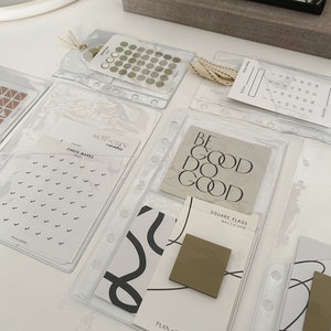 May include: A set of clear plastic pockets with various planner stickers and note cards. The stickers include a set of small circles, a set of triangles, and a set of check marks. The note cards include a card with the text "BE GOOD DO GOOD", a card with the text "SQUARE FLAGS MACCHIATO", and a card with the text "TAPE FLAGS MACCHIATO".