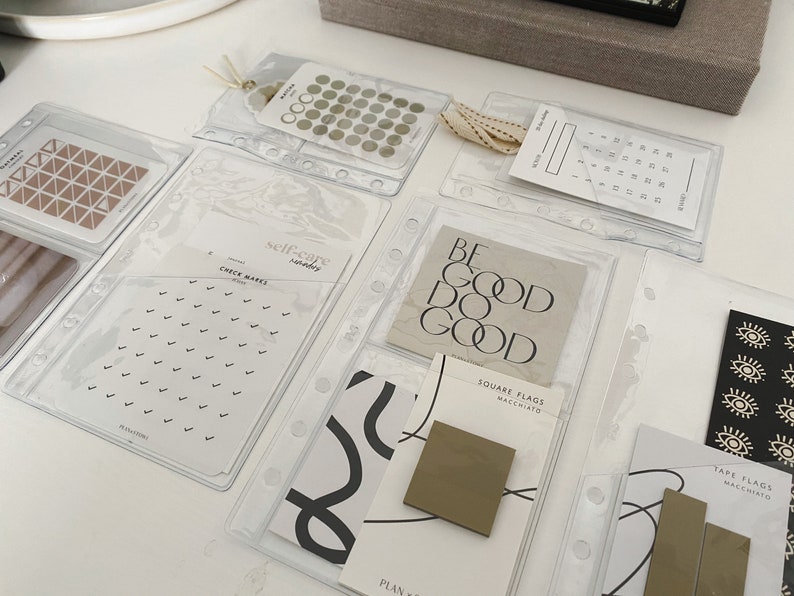 May include: A set of clear plastic pockets with various planner stickers and note cards. The stickers include a set of small circles, a set of triangles, and a set of check marks. The note cards include a card with the text "BE GOOD DO GOOD", a card with the text "SQUARE FLAGS MACCHIATO", and a card with the text "TAPE FLAGS MACCHIATO".