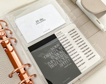 May include: A light grey planner with rose gold rings, containing inserts with the text "28 day challenge" and "Your direction is more important than your speed." A white pen with a beige grip is on the planner.