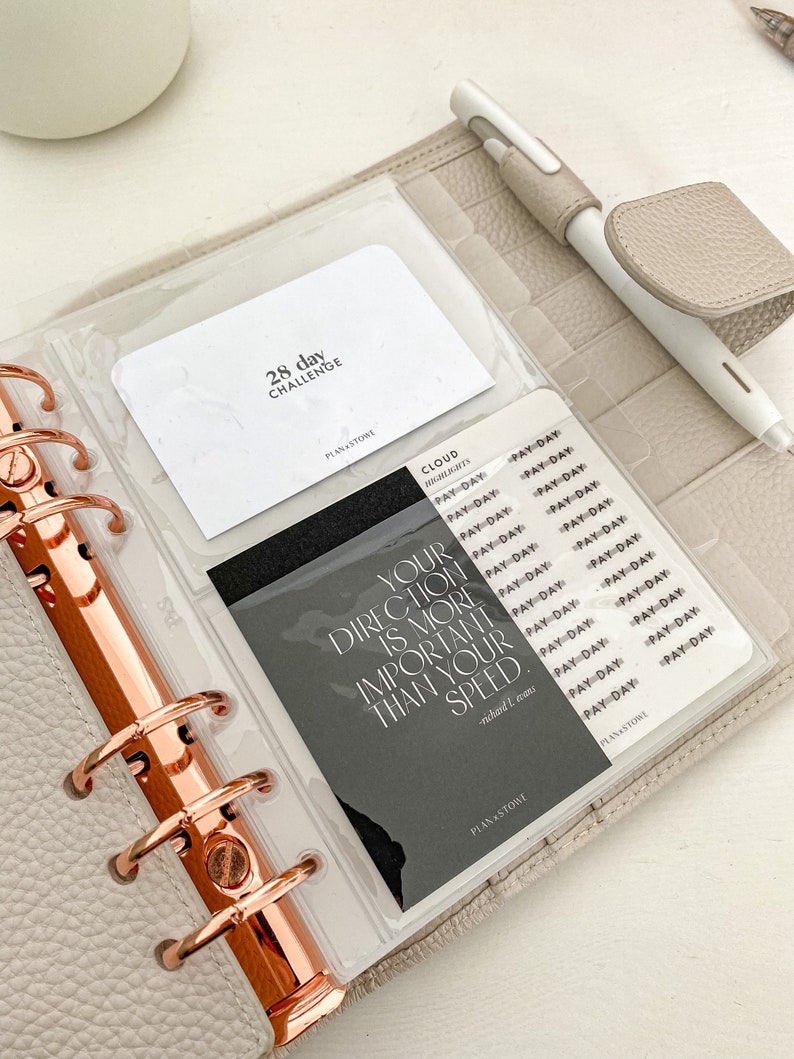 May include: A light gray planner with rose gold rings, holding inserts with text that reads "28 day challenge" and "Your direction is more important than your speed." A white pen with a beige grip rests on the planner.