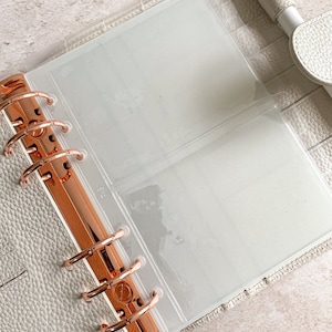 May include: An open, light gray binder with rose gold hardware and clear plastic inserts. The binder is made of textured material and has a pen loop. The binder is designed for organizing and storing documents or photos.