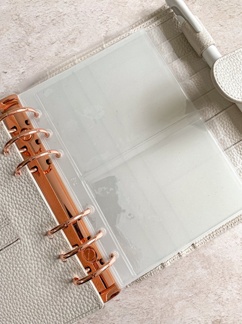May include: An open, light gray binder with rose gold hardware and clear plastic inserts. The binder is made of textured material and has a pen loop. The binder is designed for organizing and storing documents or photos.