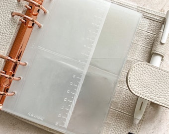 May include: A white leather planner with rose gold rings and a clear plastic pocket with a ruler. The planner has the text "PLANXSTOWE" printed on the front and the text "MOTERM" printed on the back.