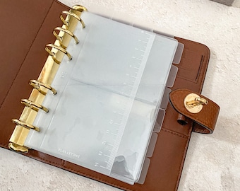 May include: A brown leather planner with gold rings and clear plastic dividers. The dividers have a ruler printed on them and the text "PLAN & STOWE" is printed on one of the dividers.