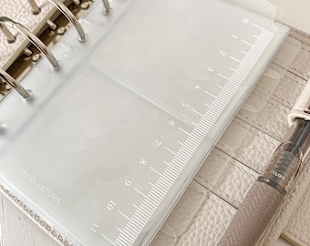 May include: Clear plastic divider with a ruler printed on the side, labeled with numbers 0 to 11. The divider is inserted into a white leather planner with silver rings.