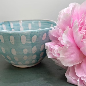 May include: A ceramic bowl with a light blue glaze and white dot pattern. A large pink peony flower is next to the bowl. The bowl is a decorative item.